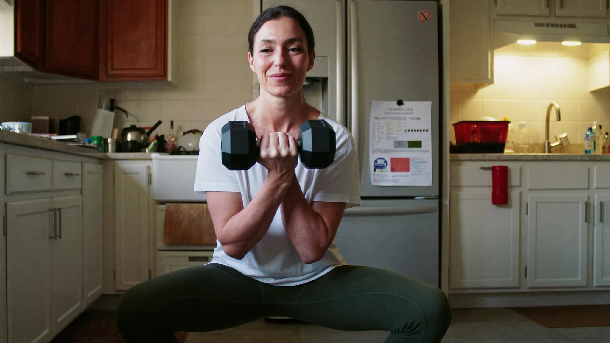 A woman in her late 40s doing a dumbbell squat in her own kitchen, the one page plan on the fridge behind her