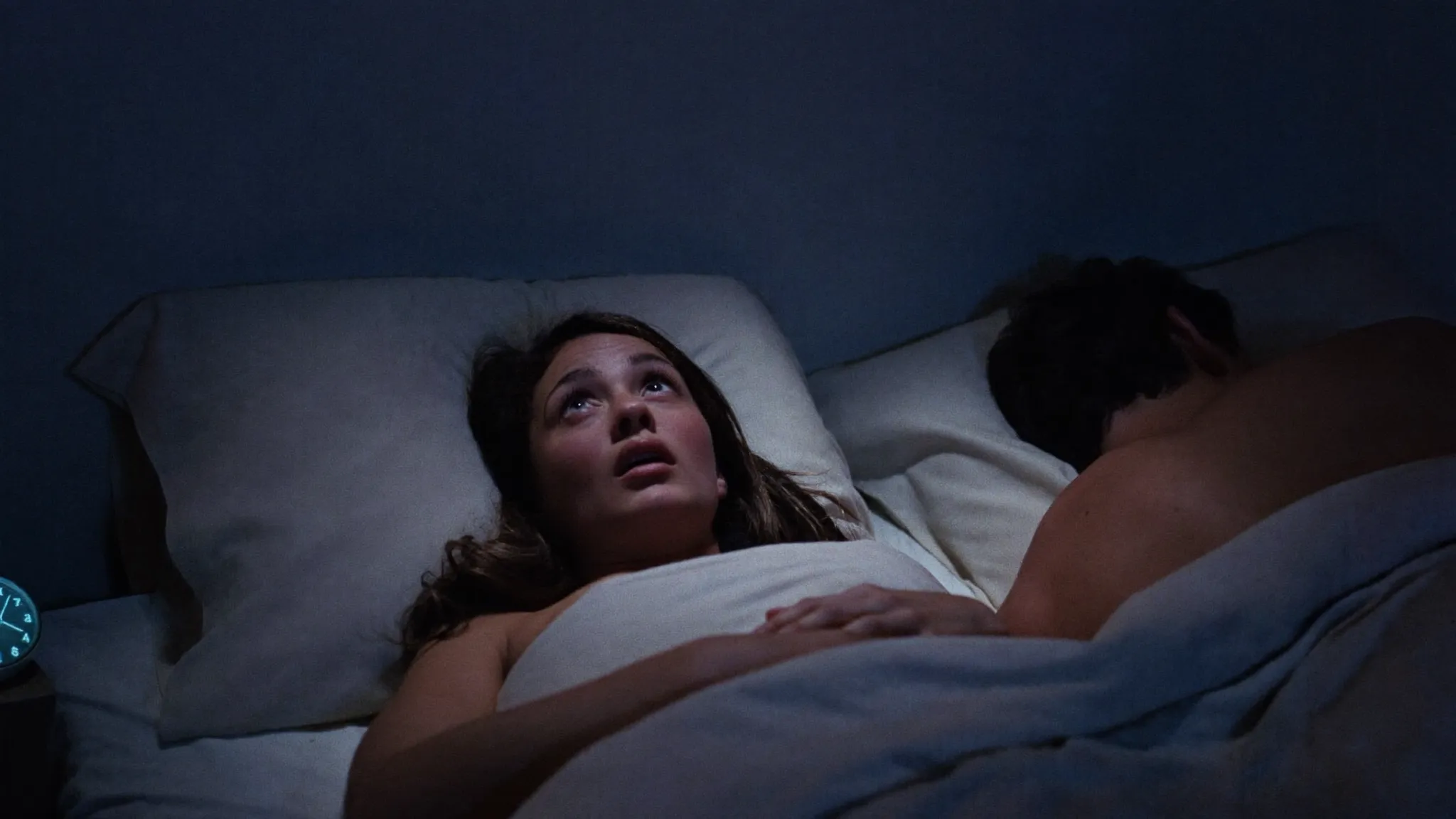 A woman lying awake in bed at night, eyes open, lit only by the bedside clock